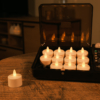 Een set van 12 oplaadbare waxinelichtjes (tot 400 uur brandduur, waterdicht, met afstandsbediening & timer) staat in een zwarte oplaadkoffer op een houten tafel voor een gezellige sfeer; één kaars zit buiten de koffer.