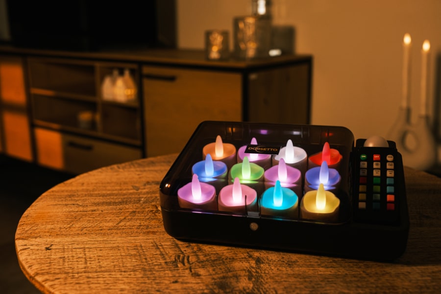 Een tafelmodel elektronisch geheugenspel met oplichtende lampjes ligt op een ronde tafel, omringd door 12 oplaadbare RGB led theelichtjes, waterdicht met afstandsbediening en timerfunctie (68 uur brandtijd), in een warm verlichte kamer met kaarsen en een kast op de achtergrond.