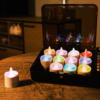 Twaalf oplaadbare, waterdichte RGB LED-theelichtjes met afstandsbediening en timer (werkingsduur van 68 uur) staan uitgestald op een houten tafel; eentje zit buiten de kist. Een gezellige, schemerig verlichte kamer vormt de achtergrond.