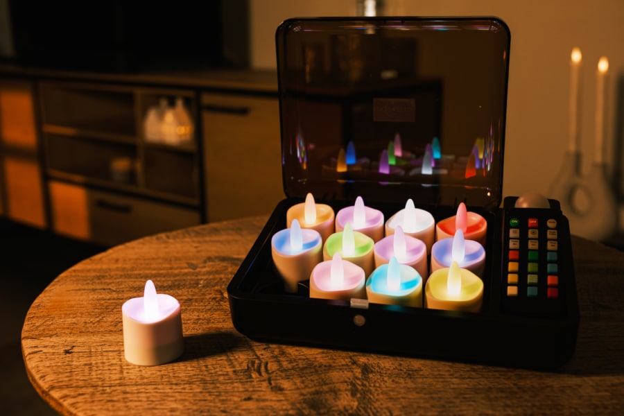 Twaalf oplaadbare, waterdichte RGB LED-theelichtjes met afstandsbediening en timer (werkingsduur van 68 uur) staan uitgestald op een houten tafel; eentje zit buiten de kist. Een gezellige, schemerig verlichte kamer vormt de achtergrond.