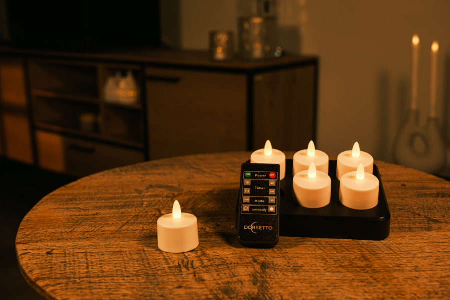 Zes dimbare, oplaadbare LED theelichtjes met een zwarte afstandsbediening (brandtijd 400 uur, waterdicht, timer) staan op een ronde houten tafel in een gezellige, warm verlichte kamer. Een kandelaar en extra kaarsen verschijnen op de achtergrond.