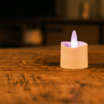 Het LED theelichtje oplaadbaar RGB LOS DORSETTO met een witte basis gloeit zacht op een houten tafel en creëert een gezellige sfeer - ideaal voor sfeerverlichting in huis.