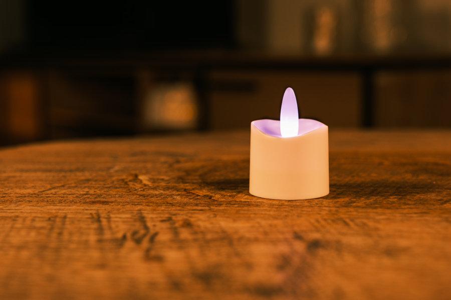 Het LED theelichtje oplaadbaar RGB LOS DORSETTO met een witte basis gloeit zacht op een houten tafel en creëert een gezellige sfeer - ideaal voor sfeerverlichting in huis.