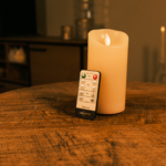 Een Oplaadbare led kaars (7,5 cm x 15 cm, 30 uur brandtijd) en de afstandsbediening staan op een ronde houten tafel, met schemerige kaarsen en een kast op de achtergrond.