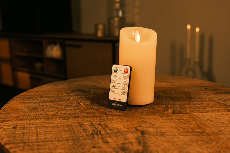 Een Oplaadbare led kaars (7,5 cm x 15 cm, 30 uur brandtijd) en de afstandsbediening staan op een ronde houten tafel, met schemerige kaarsen en een kast op de achtergrond.