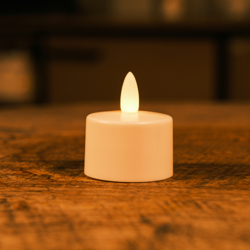 Een LED theelichtje waterdicht oplaadbaar 175 uur warm wit LOS DORSETTO met een gloeiende vlam staat op een houten oppervlak in een warm verlichte kamer.