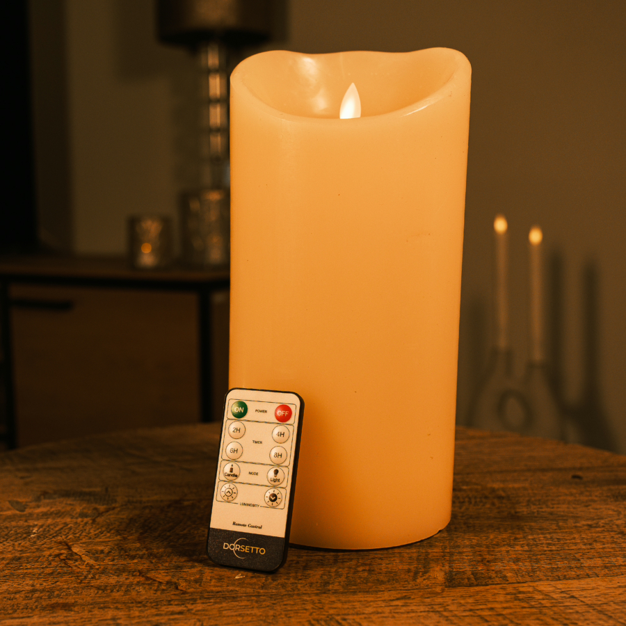 Een Oplaadbare led kaars (12,5 cm x 25 cm, 30 uur brandtijd) ligt op een houten tafel met de afstandsbediening ervoor. De achtergrond toont een schemerige kamer met meubels en kaarsen.