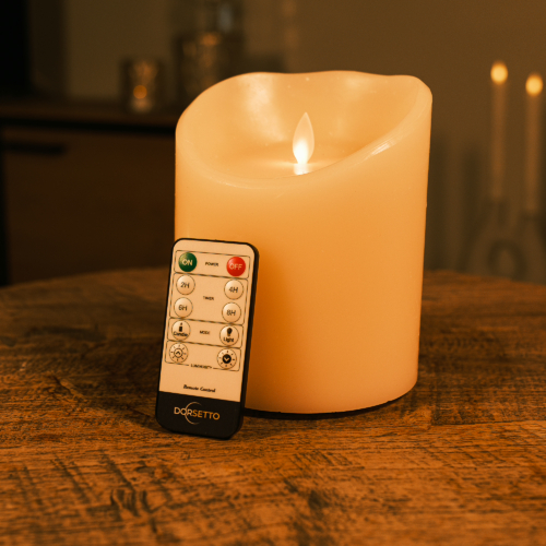 Een afstandsbediening rust tegen een Oplaadbare led kaars (12,5 cm x 15 cm, 30 uur brandtijd) met een realistisch flikkerend vlammetje, beide geplaatst op een houten tafel in een schemerige kamer.