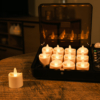 Een set van 12 led waxinelichtjes (tot 400 uur brandduur, waterdicht, kaarsvet look, met afstandsbediening) is in een laadstation op een houten tafel geplaatst; één LED-kaarsje staat er los bij. De kamer heeft warme, sfeervolle verlichting.