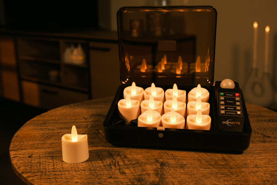 Een set van 12 led waxinelichtjes (tot 400 uur brandduur, waterdicht, kaarsvet look, met afstandsbediening) is in een laadstation op een houten tafel geplaatst; één LED-kaarsje staat er los bij. De kamer heeft warme, sfeervolle verlichting.