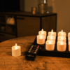 Een set van 12 oplaadbare led waxinelichtjes met afstandsbediening (tot 400 uur brandduur, waterdicht, kaarsvet look) staat op een houten tafel, met enkele kaarsen op een zwart dienblad en een gezellige kamer op de achtergrond.
