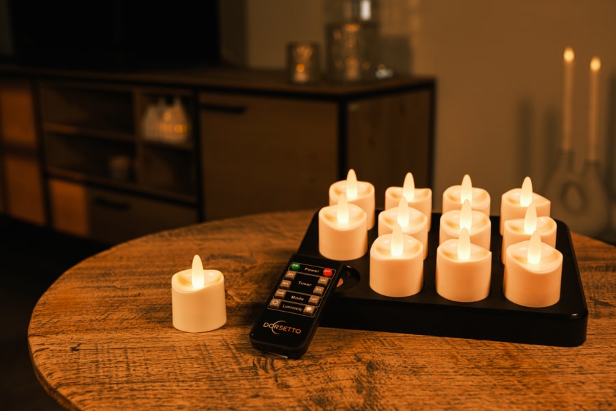 Een set van 12 oplaadbare led waxinelichtjes met afstandsbediening (tot 400 uur brandduur, waterdicht, kaarsvet look) staat op een houten tafel, met enkele kaarsen op een zwart dienblad en een gezellige kamer op de achtergrond.