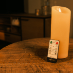 Een Oplaadbare led kaars (7,5 x 20 cm, 30 uur brandtijd) met afstandsbediening rust op een houten tafel in een warm verlichte kamer, vage meubels en kaarsen op de achtergrond.