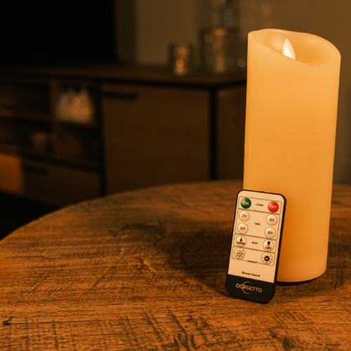 Een Oplaadbare led kaars (7,5 x 20 cm, 30 uur brandtijd) met afstandsbediening rust op een houten tafel in een warm verlichte kamer, vage meubels en kaarsen op de achtergrond.