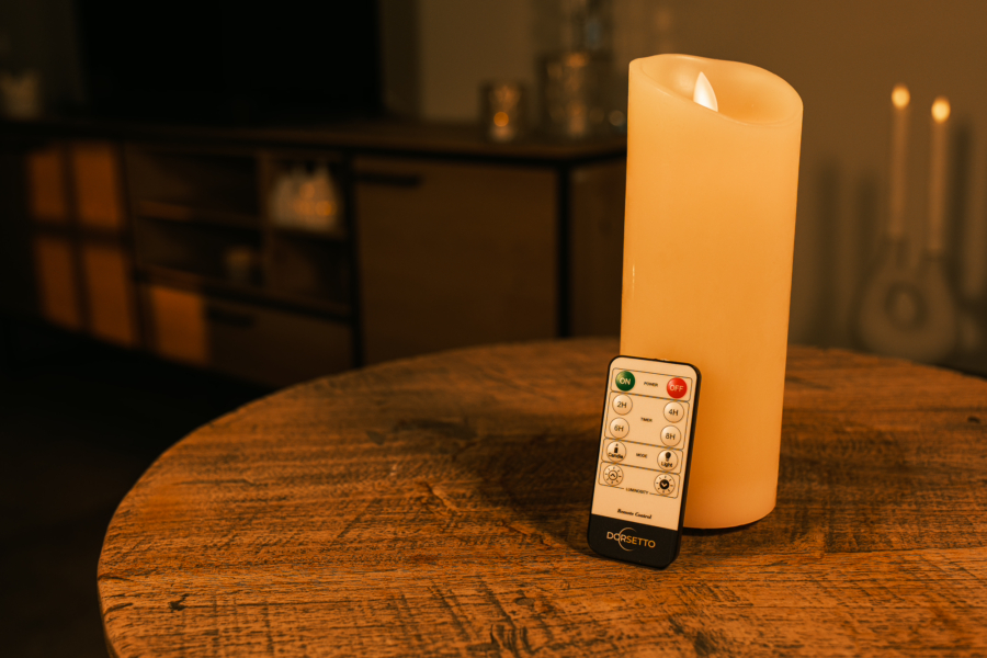 Een Oplaadbare led kaars (7,5 x 20 cm, 30 uur brandtijd) met afstandsbediening rust op een houten tafel in een warm verlichte kamer, vage meubels en kaarsen op de achtergrond.