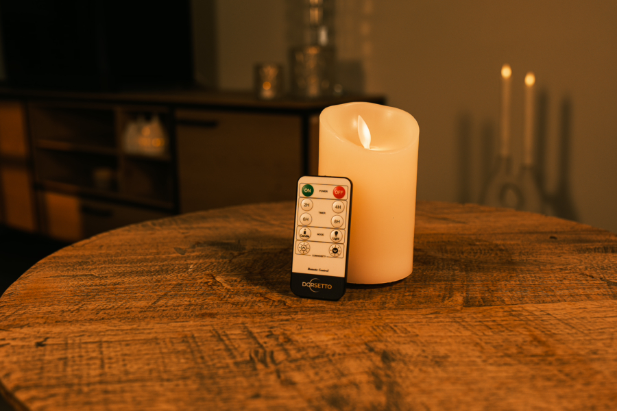 Een Oplaadbare led kaars (7,5cm x 12,5 cm, 30 uur brandtijd) met afstandsbediening staat op een ronde houten tafel in een warm verlichte kamer, met soft-focus meubels en zacht gloeiende kaarsen op de achtergrond.