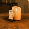 Een afstandsbediening ligt tegen een oplaadbare led kaarsen (7,5cm x 12,5cm; 30 uur brandtijd, afstandsbediening) op een houten tafel, met warme sfeerverlichting op de achtergrond.