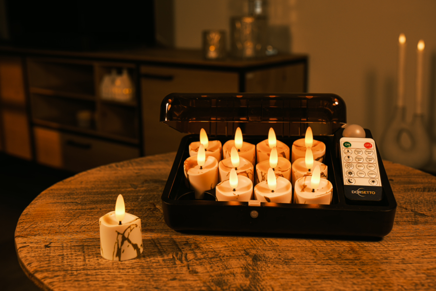 Een set van 12 oplaadbare LED theelichtjes met marmer look en afstandsbediening staat in een open zwarte box op een ronde houten tafel; één kaarsje ligt ernaast. Op de achtergrond is een zacht verlichte woonkamer zichtbaar.