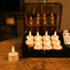 Een set van 12 oplaadbare waxinelichtjes met afstandsbediening en 3D vlam (tot 400 uur merkduur), in een zwarte opbergkist op een houten tafel; één kaars is buiten de kist geplaatst.