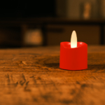 Een enkele oplaadbare rode LED theelichtje kaars met een realistische vlam (waterdicht, brandtijd 175 uur) verlicht zachtjes een houten tafel in een schemerige kamer.