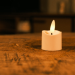 Het Realistisch led waxinelichtje oplaadbaar - los, een kleine witte led kaars met een gloeiende kunstmatige vlam, staat op een rustiek houten oppervlak tegen een zacht vervaagde achtergrond.