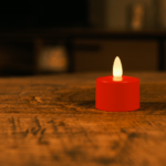 Een enkel waterdicht oplaadbaar rood LED theelichtje (brandduur 175 uur, wordt per stuk verkocht) gloeit zachtjes op een houten tafel in een zwak verlichte kamer met een onscherpe achtergrond.