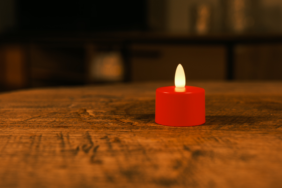Een enkel waterdicht oplaadbaar rood LED theelichtje (brandduur 175 uur, wordt per stuk verkocht) gloeit zachtjes op een houten tafel in een zwak verlichte kamer met een onscherpe achtergrond.