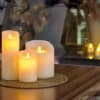 Een oplaadbare LED-kaars (7,5 x 12,5 cm) met een brandtijd van 30 uur en afstandsbediening straalt een warme gloed uit op een geweven mat bovenop een houten tafel, met vage kaarsen en decoratieve vazen op de achtergrond.