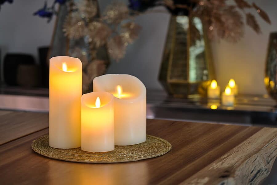 Een oplaadbare LED-kaars (7,5 x 12,5 cm) met een brandtijd van 30 uur en afstandsbediening straalt een warme gloed uit op een geweven mat bovenop een houten tafel, met vage kaarsen en decoratieve vazen op de achtergrond.