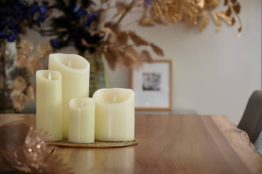 Vier Oplaadbare led kaarsen (7,5 cm x 10 cm, 30 uur brandtijd, afstandsbediening) staan op een houten tafel. De onscherpe achtergrond toont gouden en paarse bladeren met een kleine ingelijste foto aan de muur.