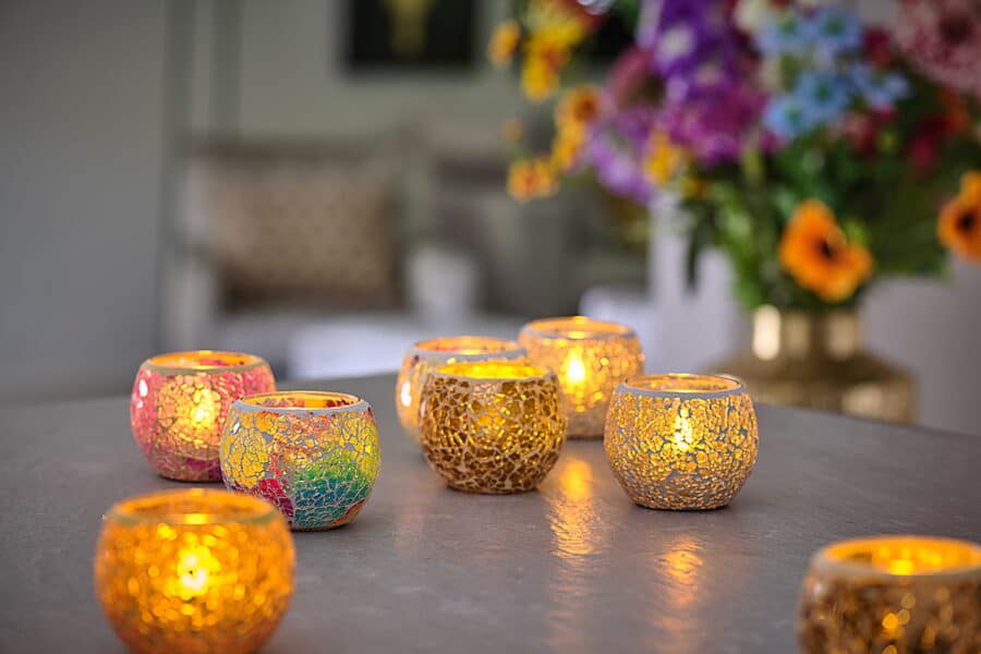 De Waxinelichthouder 8,5 CM Dorsetto mozaïek rozenkandelaars met brandende waxinelichtjes staan uitgestald op een tafel, met een vage achtergrond van levendige bloemen en een warm verlichte woonruimte.