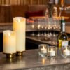 Op een warm verlicht bar- of restaurantblad staan twee Oplaadbare led kaarsen (7,5cm x 12,5cm, 30u brandtijd, afstandsbediening), drie votiefkaarsen, wijnflessen, glaswerk en kleine vaasjes met bloemen.