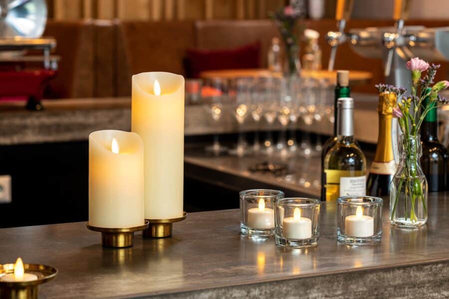 Op een warm verlicht bar- of restaurantblad staan twee Oplaadbare led kaarsen (7,5cm x 12,5cm, 30u brandtijd, afstandsbediening), drie votiefkaarsen, wijnflessen, glaswerk en kleine vaasjes met bloemen.