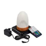 Een compacte, ovaalvormige oplaadbare led tafellamp in marmerwit met een voet in houtlook staat op een zwart plateau, samen met een zwarte voedingsadapter en een kleurrijke afstandsbediening. De lamp is waterdicht en heeft een timerfunctie.