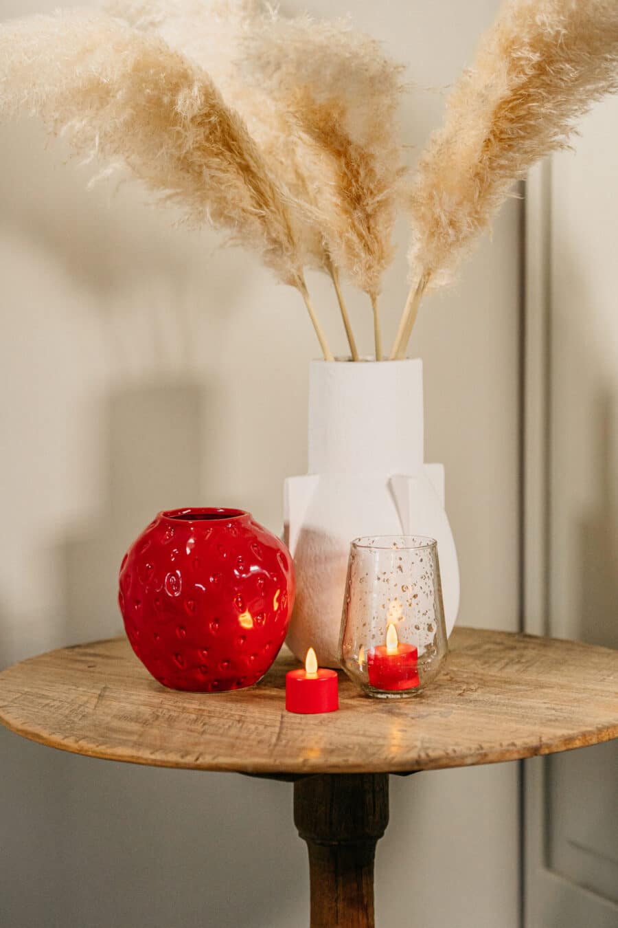 Op een ronde houten tafel staan een witte vaas met pampagras, een rode vaas met structuur, een helder glas met gouden spikkels en twee rode oplaadbare waxinelichtjes uit de set van 12 (waterdicht, afstandsbediening, 175 uur batterijduur).