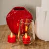 Een rode glazen vaas, een wit object van keramiek en twee rode oplaadbare waxinelichtjes (waterdicht, met afstandsbediening, 175 uur brandtijd) - een in getextureerd glas en een daarbuiten - staan op een houten oppervlak.
