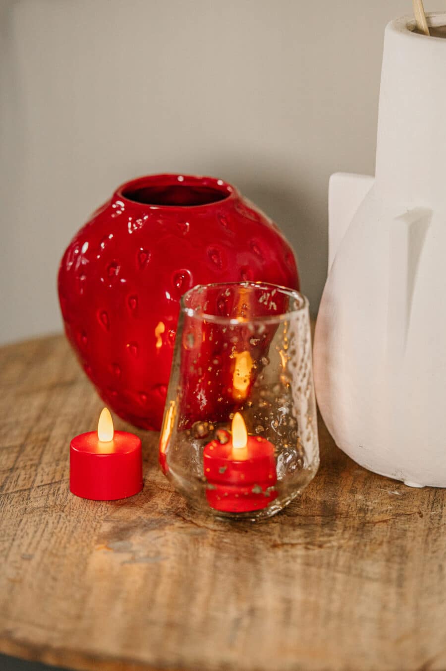 Een rode glazen vaas, een wit object van keramiek en twee rode oplaadbare waxinelichtjes (waterdicht, met afstandsbediening, 175 uur brandtijd) - een in getextureerd glas en een daarbuiten - staan op een houten oppervlak.