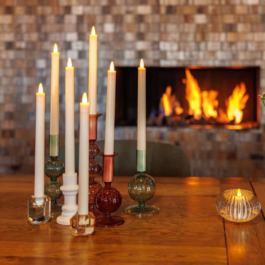 Een set van 12 oplaadbare dinerkaarsen (270mm, warm wit, 130 uur brandtijd, met afstandsbediening) is gerangschikt op een houten tafel, naast een waxinelichtje en voor een gloeiende open haard.