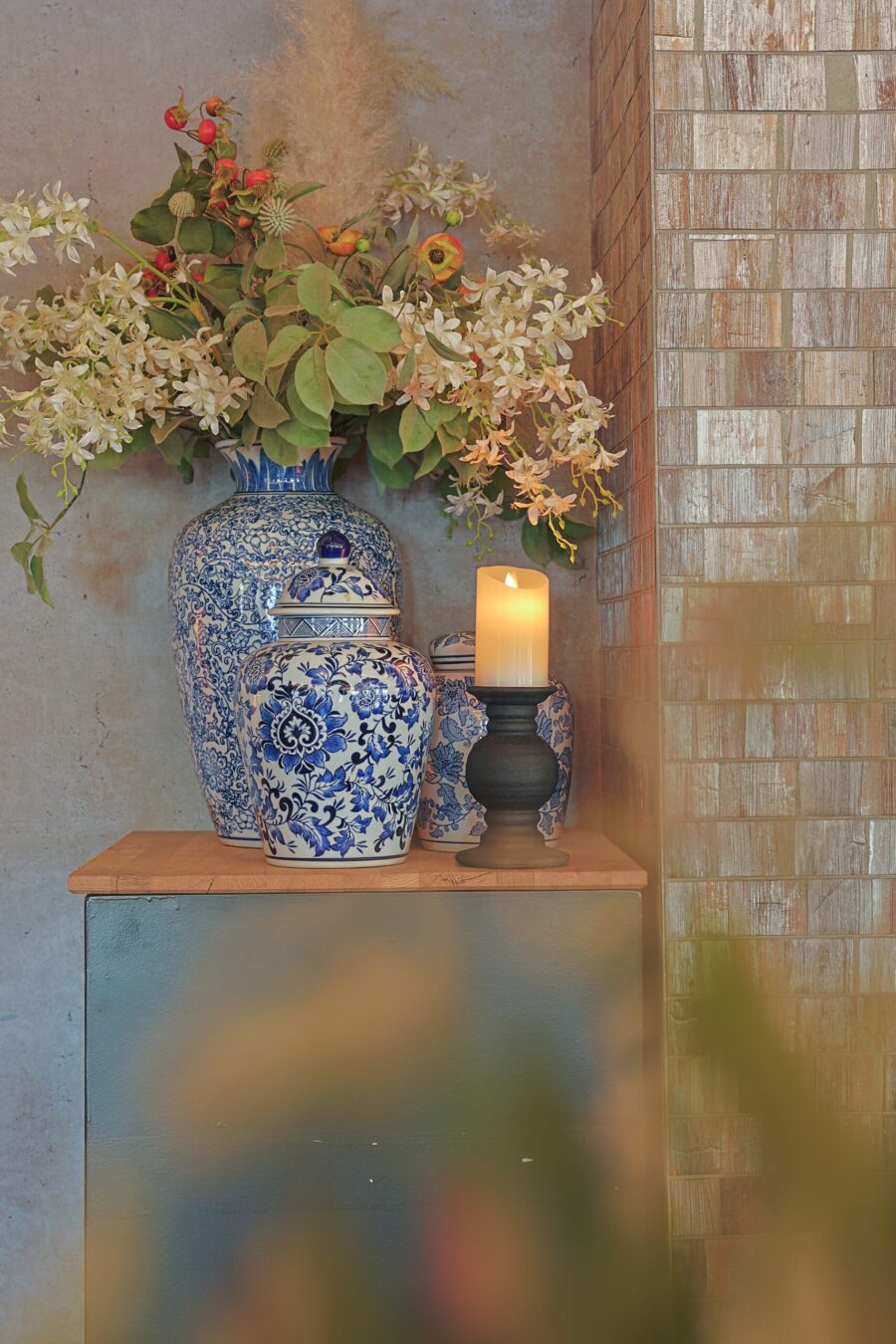 Blauwe en witte keramieken vazen met bloemmotieven bevatten witte bloemen en groen op een houten sokkel. Daarnaast voegt een Oplaadbare led kaars (30 uur brandtijd, afstandsbediening, 7,5x10 cm) in een zwarte houder warmte toe tegen de muur met textuur.