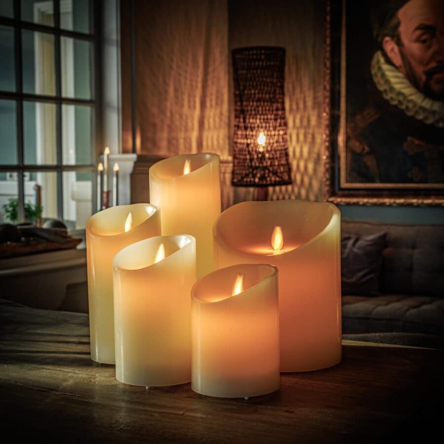 Een groep van vijf Oplaadbare led kaarsen (7,5 cm x 15 cm, 30 uur brandtijd, met afstandsbediening) gloeit warm op een houten tafel in een gezellige woonkamer met een portret en groot raam op de achtergrond.