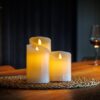 Drie oplaadbare led kaarsen (set van 3, 30 uur brandtijd, met afstandsbediening en timerfunctie) staan op een geweven mat op een houten tafel. Op de achtergrond zie je een glas rode wijn en een fles, voor een gezellige sfeer.