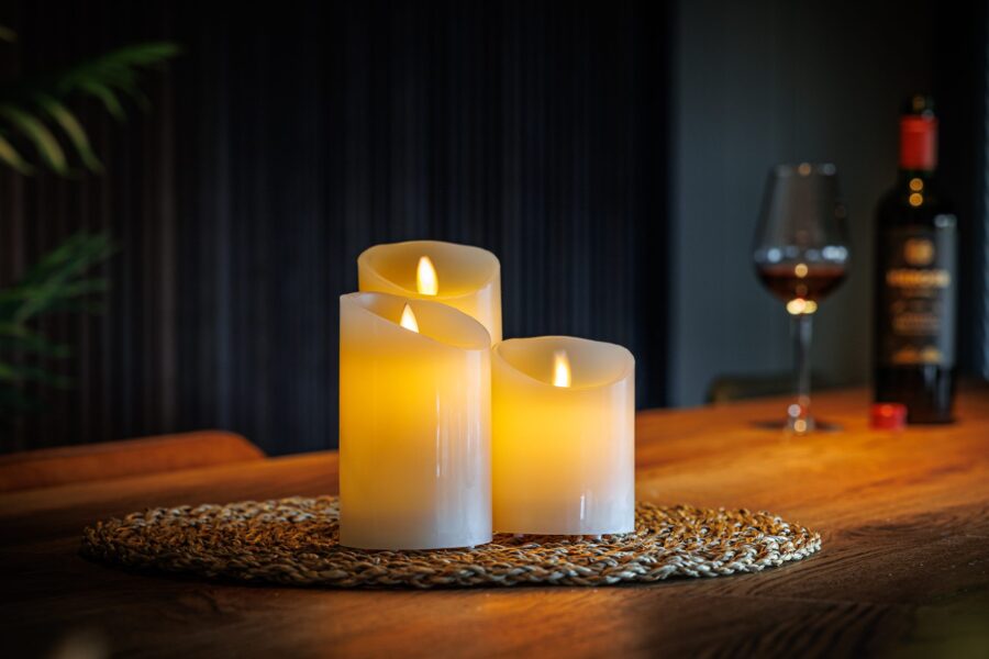 Drie oplaadbare led kaarsen (set van 3, 30 uur brandtijd, met afstandsbediening en timerfunctie) staan op een geweven mat op een houten tafel. Op de achtergrond zie je een glas rode wijn en een fles, voor een gezellige sfeer.