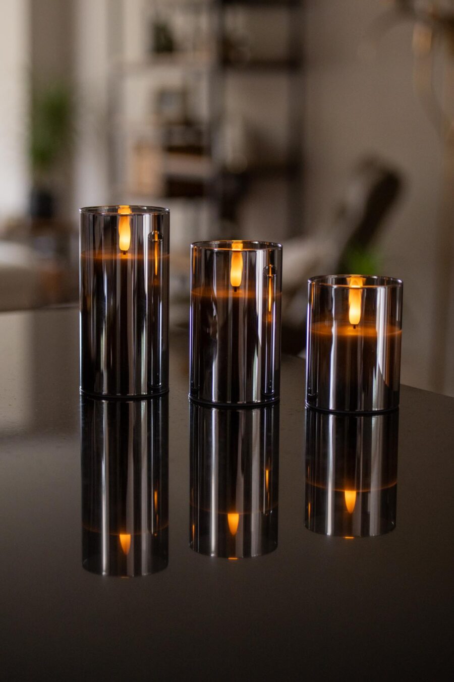 Drie donkergrijze oplaadbare led kaarsen (set van 3), met afstandsbediening en 105 uur brandtijd, staan op een glanzend zwart oppervlak en verspreiden warme weerspiegelingen in een gezellig, modern interieur.