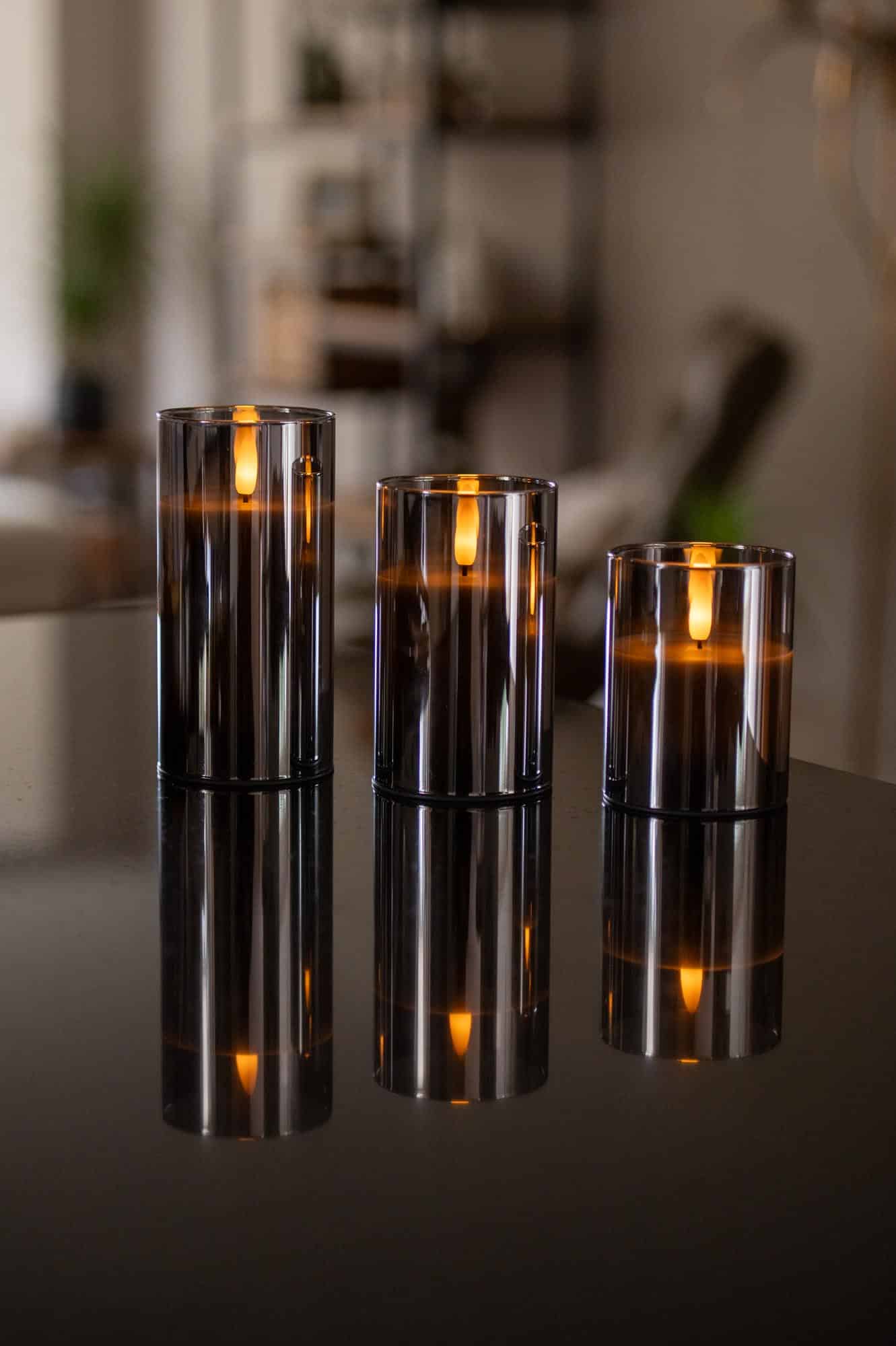 Drie donkergrijze oplaadbare led kaarsen (set van 3), met afstandsbediening en 105 uur brandtijd, staan op een glanzend zwart oppervlak en verspreiden warme weerspiegelingen in een gezellig, modern interieur.