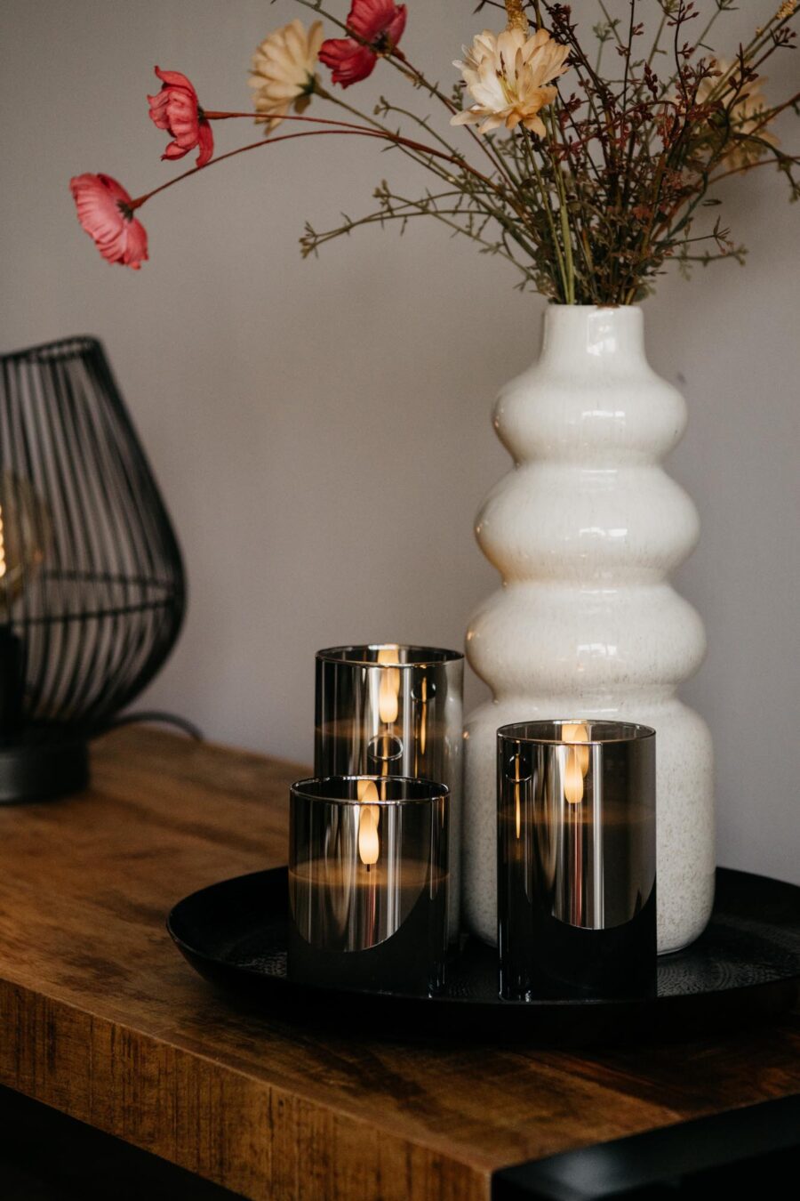 Een witte vaas van keramiek met rode en crèmekleurige bloemen staat op een houten tafel naast drie donkergrijze oplaadbare ledkaarsen (3 oplaadbare ledkaarsen, met afstandsbediening, 105 uur brandtijd) gerangschikt op een rond zwart dienblad.