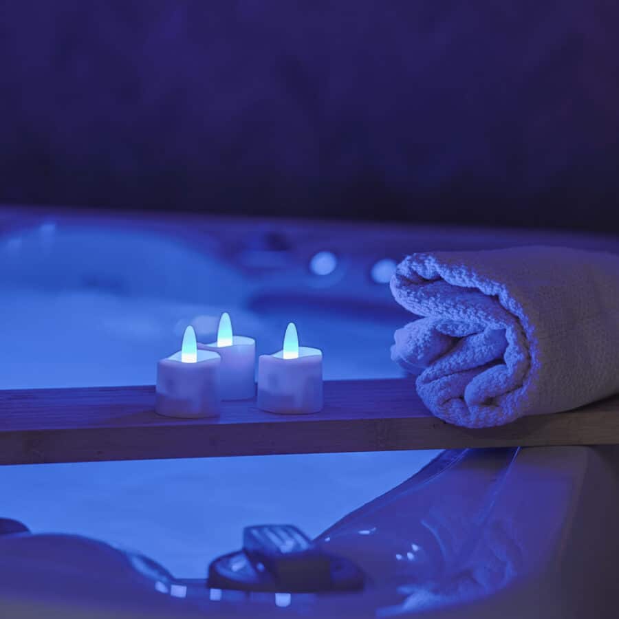 Een ontspannend tafereel met opgerolde handdoeken en zes RGB oplaadbare waxinelichtjes (waterdicht, 68 uur brandtijd, met afstandsbediening en timerfunctie) op een houten dienblad bij een zacht verlicht bad voor een serene spa-achtige sfeer.