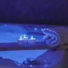 Een opgerolde witte handdoek en drie oplaadbare RGB led theelichtjes (waterdicht, met afstandsbediening en timer, 68 uur brandtijd) zitten op een houten dienblad boven een zacht verlichte blauwe jacuzzi en creëren een ontspannen spasfeer.