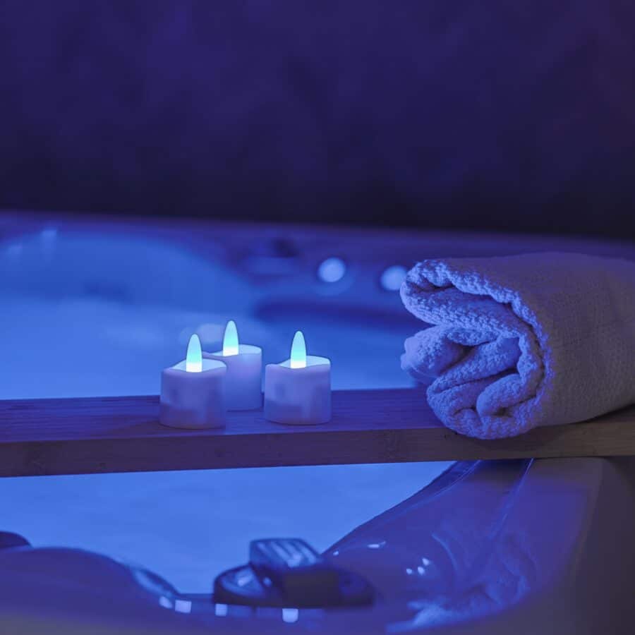 Een opgerolde witte handdoek en drie oplaadbare RGB led theelichtjes (waterdicht, met afstandsbediening en timer, 68 uur brandtijd) zitten op een houten dienblad boven een zacht verlichte blauwe jacuzzi en creëren een ontspannen spasfeer.