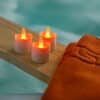 Drie oplaadbare RGB LED theelichtjes (waterdicht, met afstandsbediening en timer, 68 uur brandtijd) uit een set van 12 staan op een houten plank naast een oranje handdoek in een spa-achtige setting met blauwe achtergrond.
