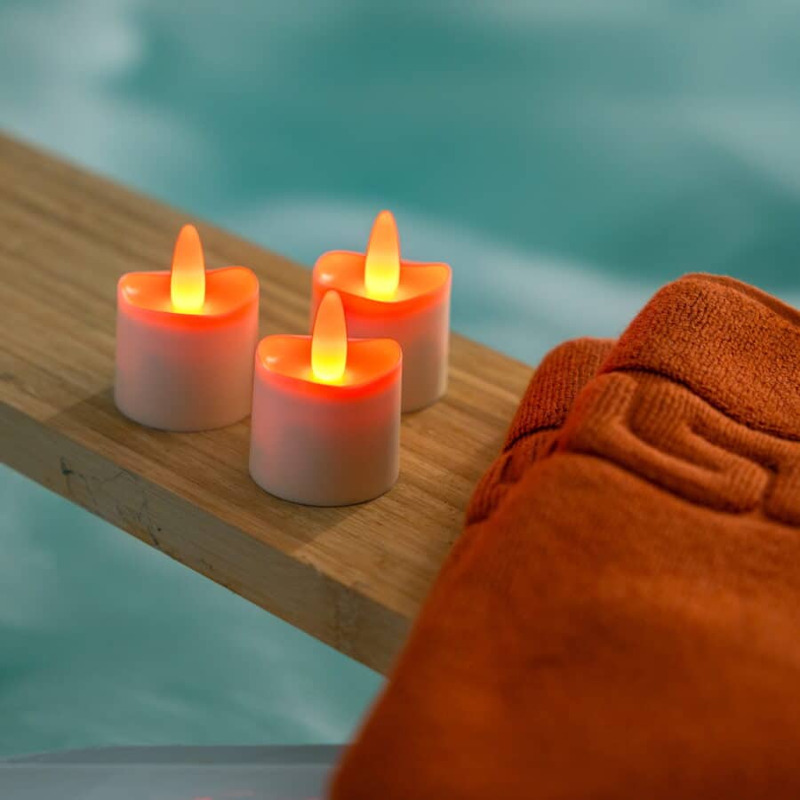 Drie oplaadbare RGB LED theelichtjes (waterdicht, met afstandsbediening en timer, 68 uur brandtijd) uit een set van 12 staan op een houten plank naast een oranje handdoek in een spa-achtige setting met blauwe achtergrond.